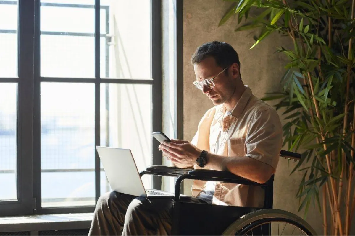 تظهر الصورة رجل من ذوي الإعاقة يمارس العمل باستخدام اللابتوب والهاتف الجوال