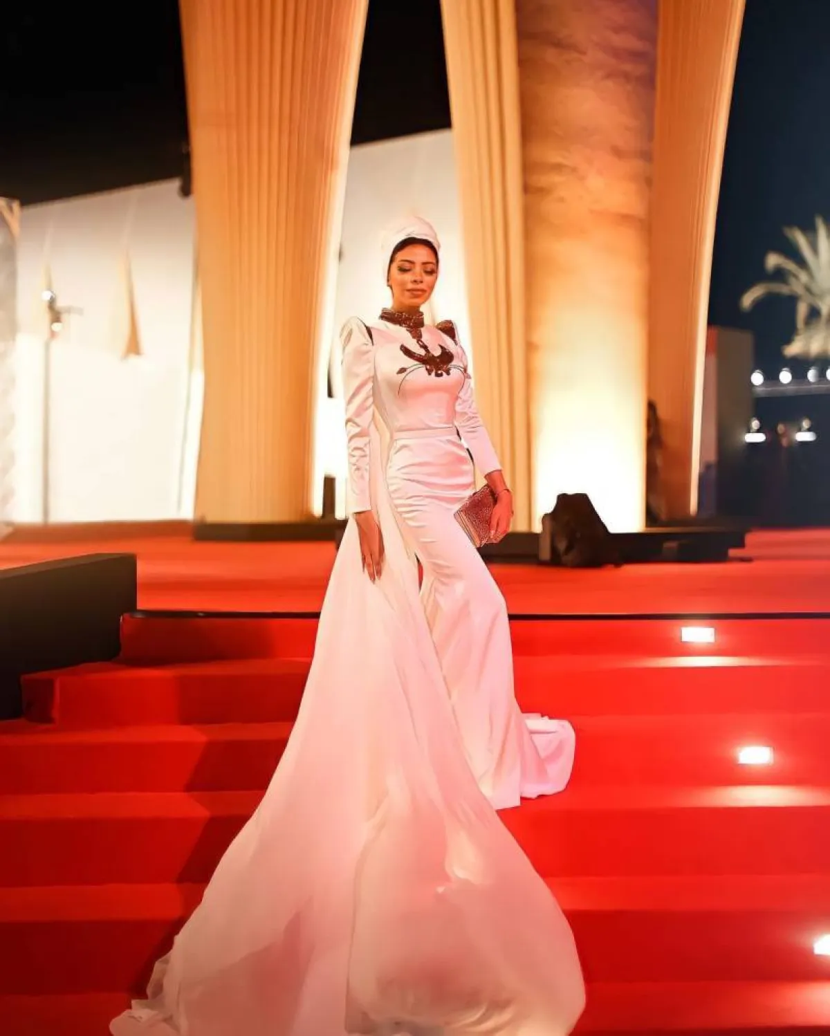 منة السُني بفستان باللون الأبيض والذهبي مع حقيبة كلاتش مستطيلة