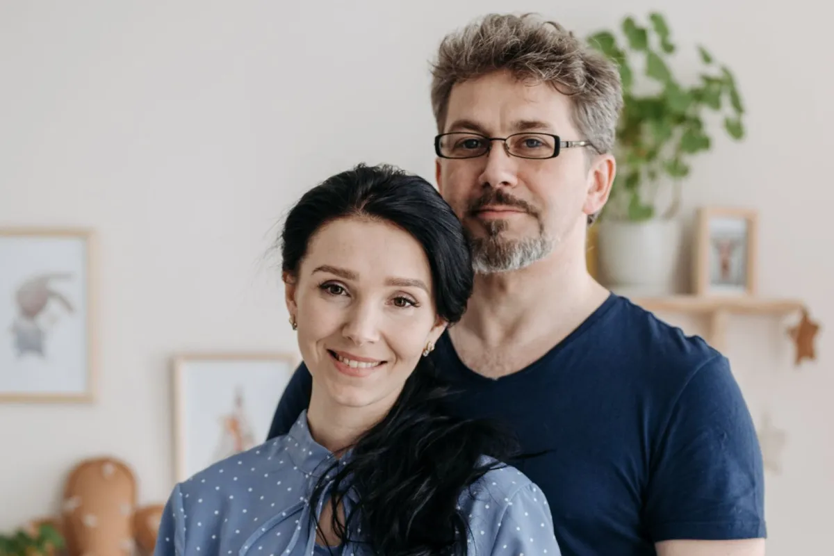 زوج يقف وراء زوجته وهما بالمنزل تبدو على ملامحه أسارير السعادة