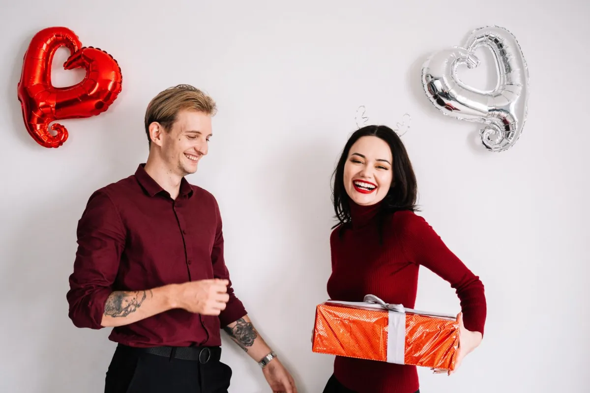 زوجة شابة تبتسم بسعادة وهي تهدي زوجها الحبيب هدية تحيط بهما بالونات من القلوب 
