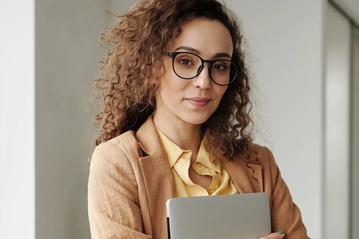 فتاة تبدو واثقة من نفسها تبتسم وهي ترتدي نظارة وتمسك بيدها جهاز كومبيوتر محمولاً  