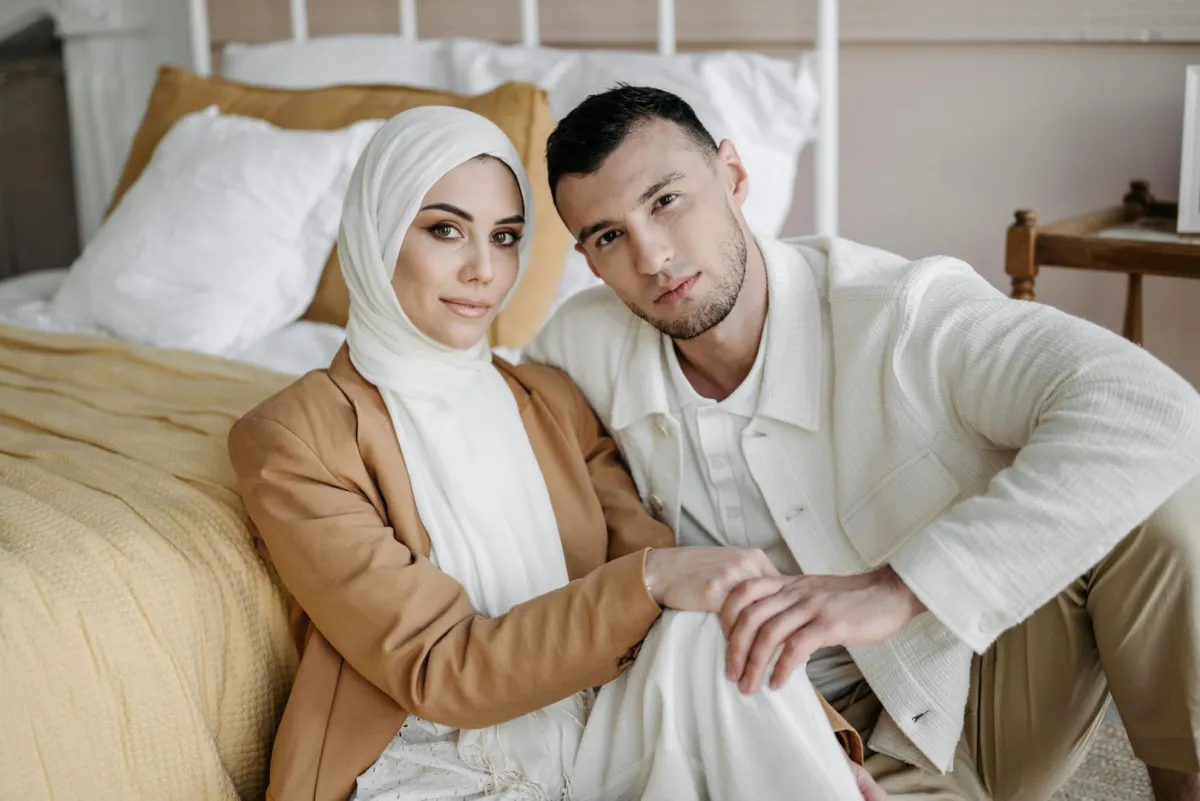 زوج يمسك يد زوجته وهما يجلسان على الأرض بجوار السرير بحجرة النوم ويبدوان سعيدين