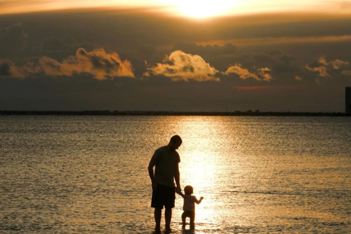 والد يأخذ بيد صغيره ليعلمه المشي بين مياه البحر الساكنة