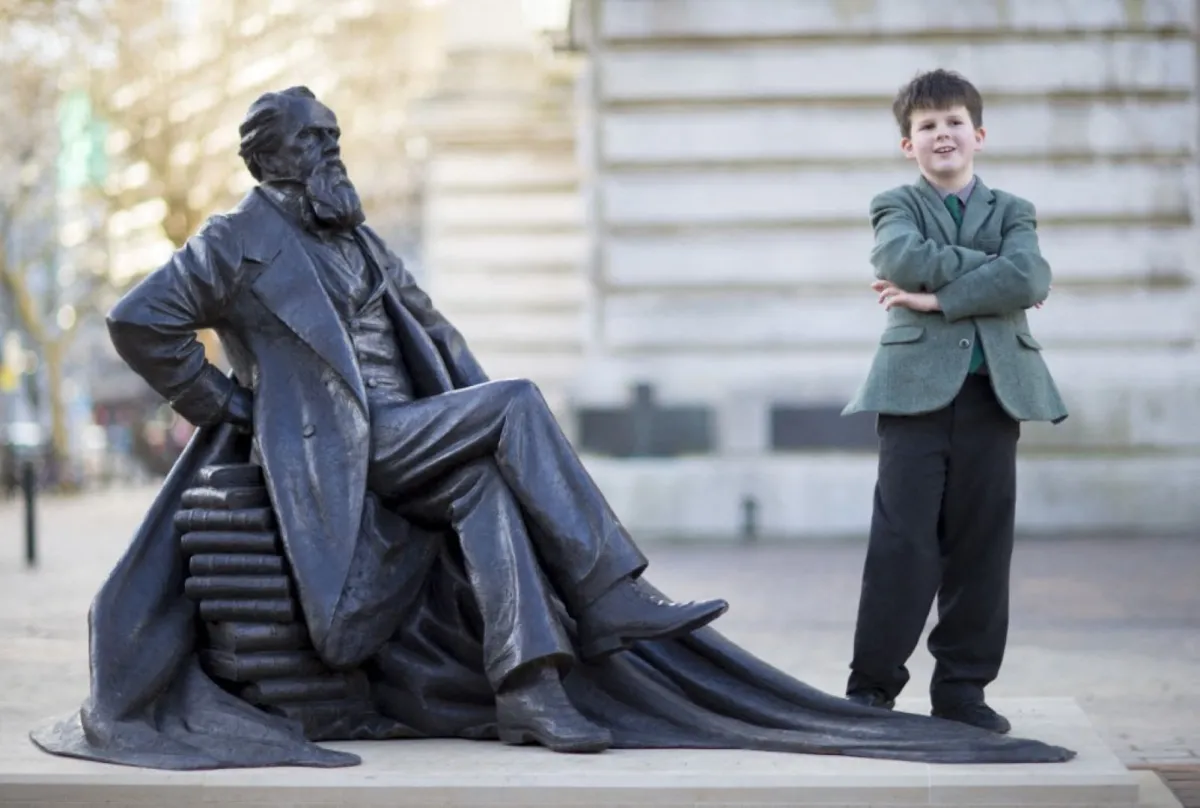 يقف أوليفر ديكنز، البالغ من العمر 9 سنوات جانب تمثال جده الأكبر، الكاتب البريطاني الراحل تشارلز ديكنز، الذي نُصب حديثاً في ساحة جيلدهول في بورتسموث، في 7 من فبراير 2014 -مصدر الصورة   CHRIS ISON / AFP