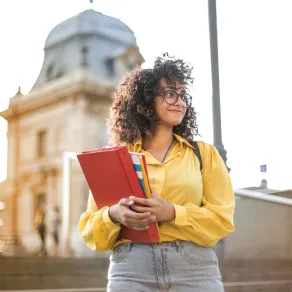 فتاة تقف أمام مبنى الجامعة وتمسك بيدها كتاباً دراسياً وهي تبتسم بسعادة مع بداية العام الدراسي الجديد