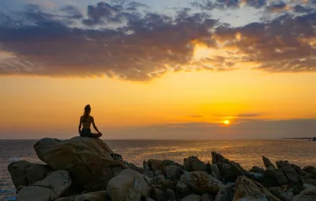 شابة تجلس على صخرة تلعب اليوغا في هدوء وتتابع قرص الشمس بين الآفاق 