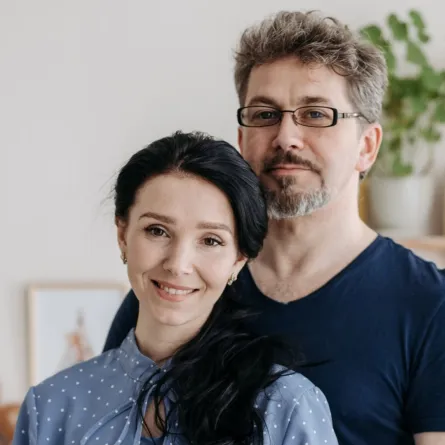 زوج يقف وراء زوجته وهما بالمنزل تبدو على ملامحه أسارير السعادة