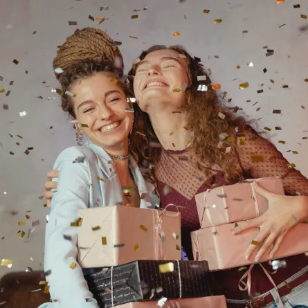 فتاة تهدي صديقتها عدداً من الهدايا وهما في قمة السعادة وتتساقط حولهما الوريقات الملونة