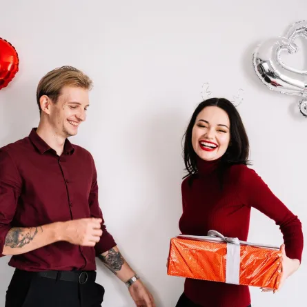 زوجة شابة تبتسم بسعادة وهي تهدي زوجها الحبيب هدية تحيط بهما بالونات من القلوب 