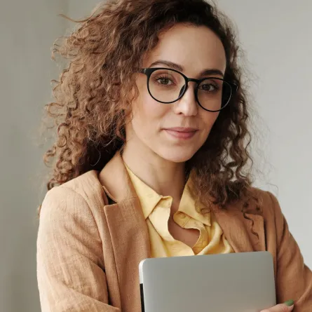 فتاة تبدو واثقة من نفسها تبتسم وهي ترتدي نظارة وتمسك بيدها جهاز كومبيوتر محمولاً  