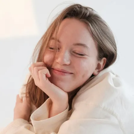 فتاة تبتسم بكسل وهي ترتدي رداء النوم وتظهر أشعة الشمس منعكسة على أحد الجدران