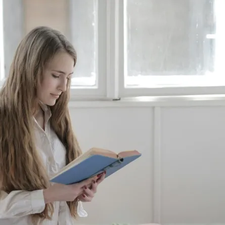 فتاة تقرأ رواية في حجرة بيضاء وبجوارها نافذة مفتوحة