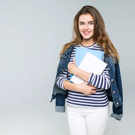 طالبة بالجامعة تحمل كتباً ومحاضرات وتبتسم بسعادة قبل دخول المحاضرة