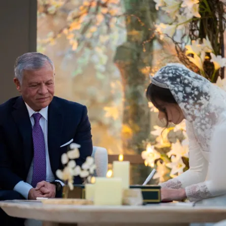 الملك عبد الله الثاني ينظر إلى ابنته الكبرى الأميرة إيمان وهي توقع وثيقة زواجها خلال حفل زفافها على جميل ألكساندر ترميوتيس في عمان (King Abdullah II looking on as his eldest daughter Princess Iman signs her marriage document during her wedding ceremony with Jameel Alexander Thermiotis in Amman). مصدر الصورة: Jordanian Royal Palace / AFP