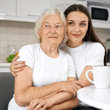 زوجة تحتضن كتف حماتها وهما جالستان بالمطبخ وأمامهما كوبان من النسكافيه