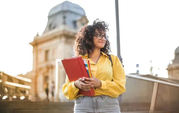 فتاة تقف أمام مبنى الجامعة وتمسك بيدها كتاباً دراسياً وهي تبتسم بسعادة مع بداية العام الدراسي الجديد
