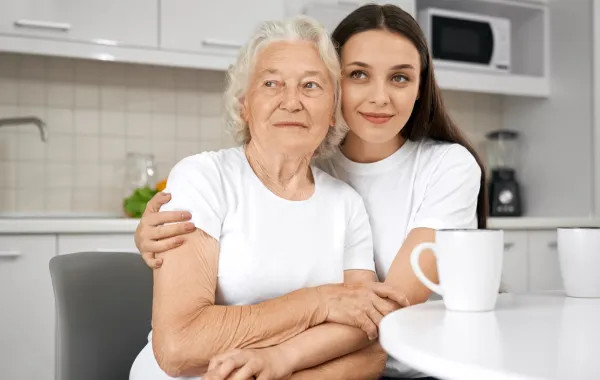 زوجة تحتضن كتف حماتها وهما جالستان بالمطبخ وأمامهما كوبان من النسكافيه