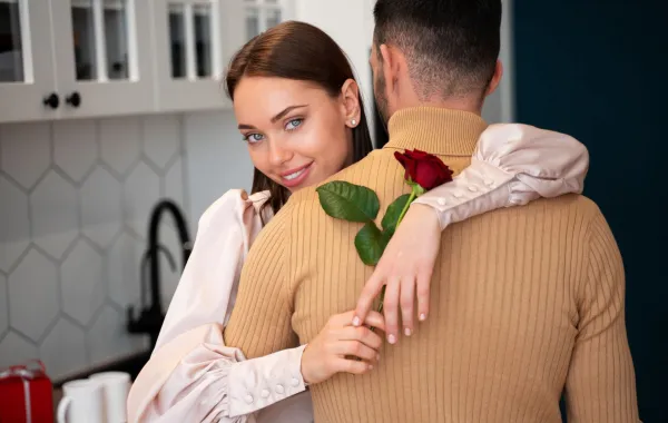 زوجة تحتضن زوجها وهي تمسك زهرة وتبتسم بدلال