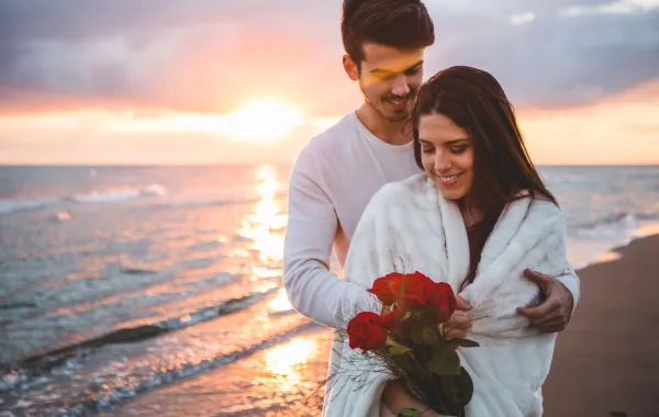 زوج يحتضن كتف زوجته وهي تبتسم بسعادة تحمل معها باقة من الزهور الحمراء على شاطئ البحر