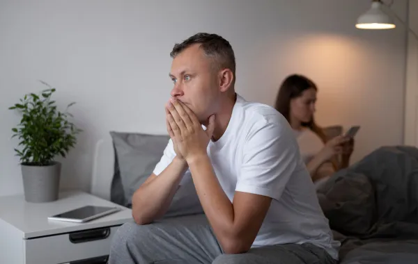 زوجة تمسك الموبايل تتحدث فيه وزوجها يجلس صامتاً يضع يديه أسفل وجهه يفكر 